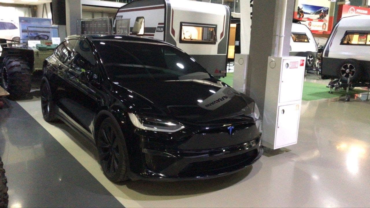 На фотографии изображен черный электромобиль Tesla Model X в интерьере большого выставочного зала. На заднем плане видны туристические автодома (рейкары) и крупное колесо вездехода, что указывает на профиль заведения. Это фото сделано не в торговом центре, а в шоуруме, специализирующемся на внедорожной технике и оборудовании для кемпинга.